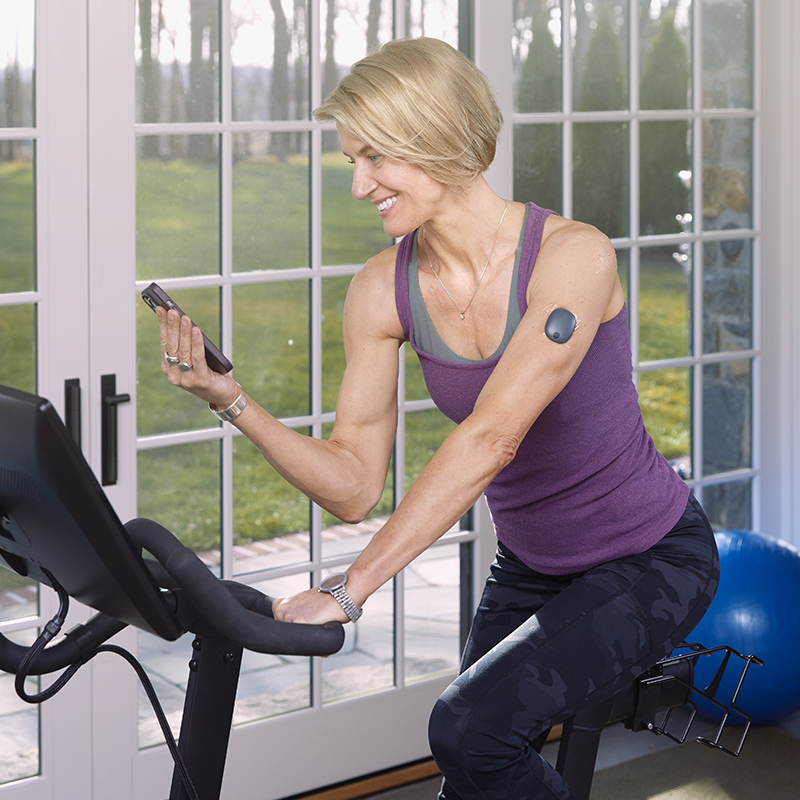 Donna in sella a una cyclette mentre utilizza un’applicazione per il monitoraggio del glucosio.