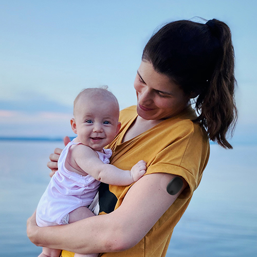 Donna che indossa un CGM Eversense a lungo termine e tiene in braccio il suo bambino sulla spiaggia.